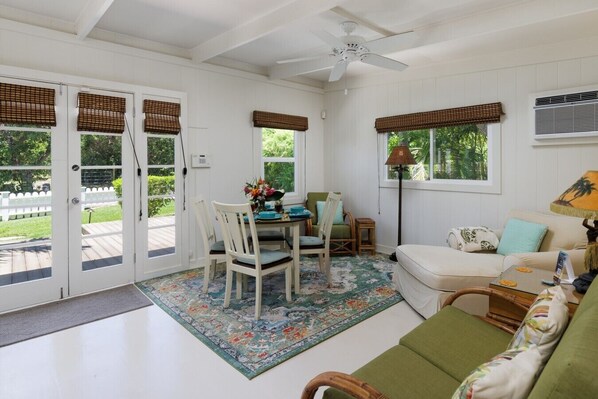A room with a dining table, chairs, a sofa, a ceiling fan, and a window with a view of greenery. - Mele Mele  3BR Puako Beach Retreat Steps to Ocean Access (Waimea)