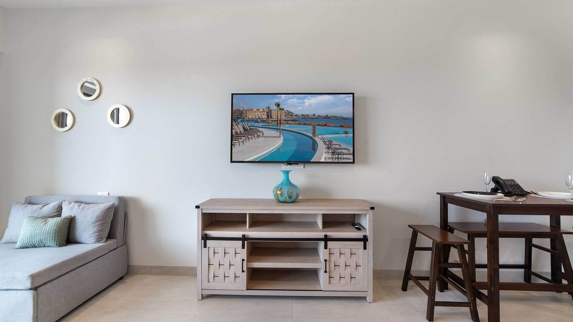 Ein Wohnzimmer mit einer Couch, einem Couchtisch mit einer Vase, einem an der Wand montierten Fernseher und einem Esstisch mit Stühlen.