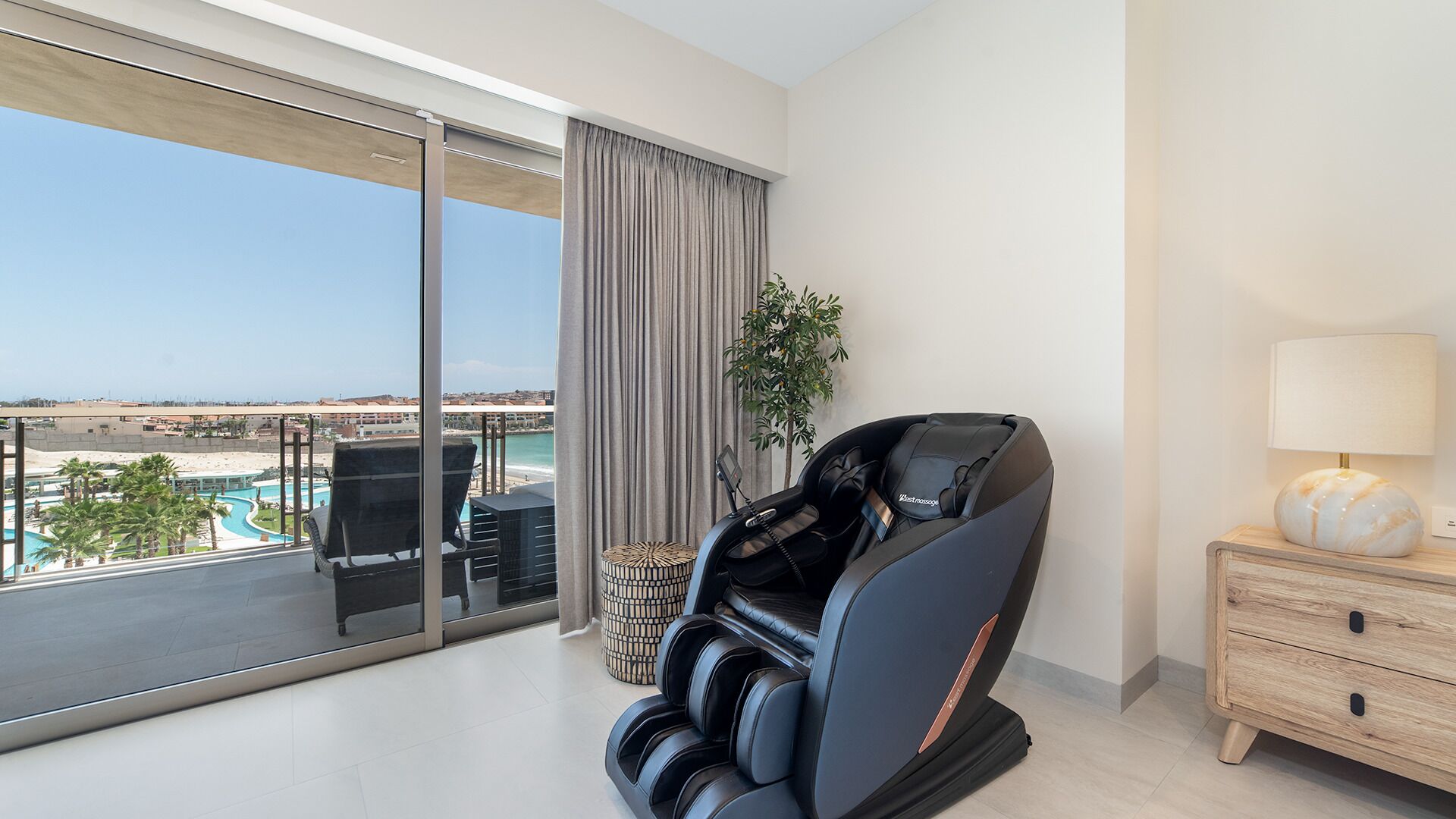 Ein modernes Hotelzimmer mit Massagesessel, Nachttisch mit Lampe und Blick auf Strand und Pool.