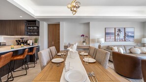 Ein moderner Essbereich mit einem langen Tisch für vier Personen, eine Küche mit dunklen Holzschränken und ein Wohnbereich mit einer Couch und dekorativen Kissen.