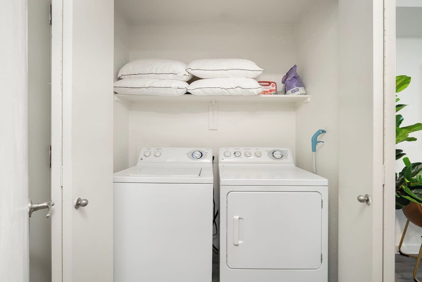 A laundry room with two white dryers, a shelf with pillows and a bag, and a blue towel rack.
