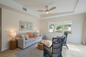 Un salon comprenant un canapé, une table basse et des fauteuils, un ventilateur de plafond et une fenêtre donnant sur un paysage verdoyant.