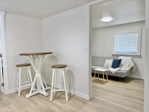 Um quarto com parede branca, piso de madeira, uma mesa branca com dois bancos e um sofá cinza com manta.