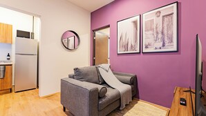 A compact living space with a kitchenette, a gray sofa, a round mirror, and two framed artworks on a purple wall.