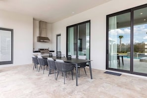 Un espace repas extérieur moderne, comprenant une table et des chaises, un barbecue et une porte vitrée donnant accès à une piscine.