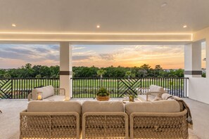 A covered outdoor seating area with a view of a green landscape and a sunset. - The Dream Mansion | Luxe 10BR w/ Private Pool, Spa, Sauna, Theater Near Disney (Kissimmee)