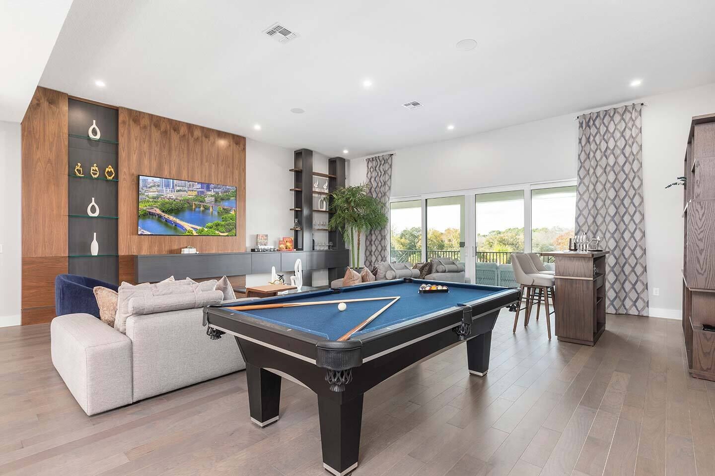 A modern living room with a pool table, comfortable seating, a TV mounted on the wall, and a balcony with a view.