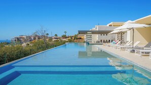 Ein klares, blaues Schwimmbecken mit Liegestühlen und Sonnenschirmen, umgeben von Gebäuden und Blick aufs Meer.
