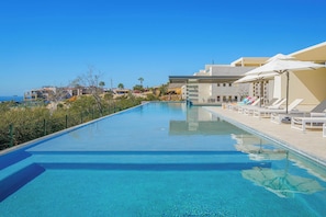 Une piscine d’un bleu éclatant, bordée de chaises longues et d’ombrelles, entourée de bâtiments et offrant une vue sur l’océan.