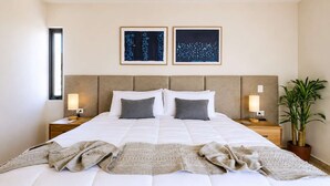 Ein Hotelzimmer mit einem großen Bett, zwei Nachttischen mit Lampen, einer Topfpflanze und zwei gerahmten Kunstwerken an der Wand.
