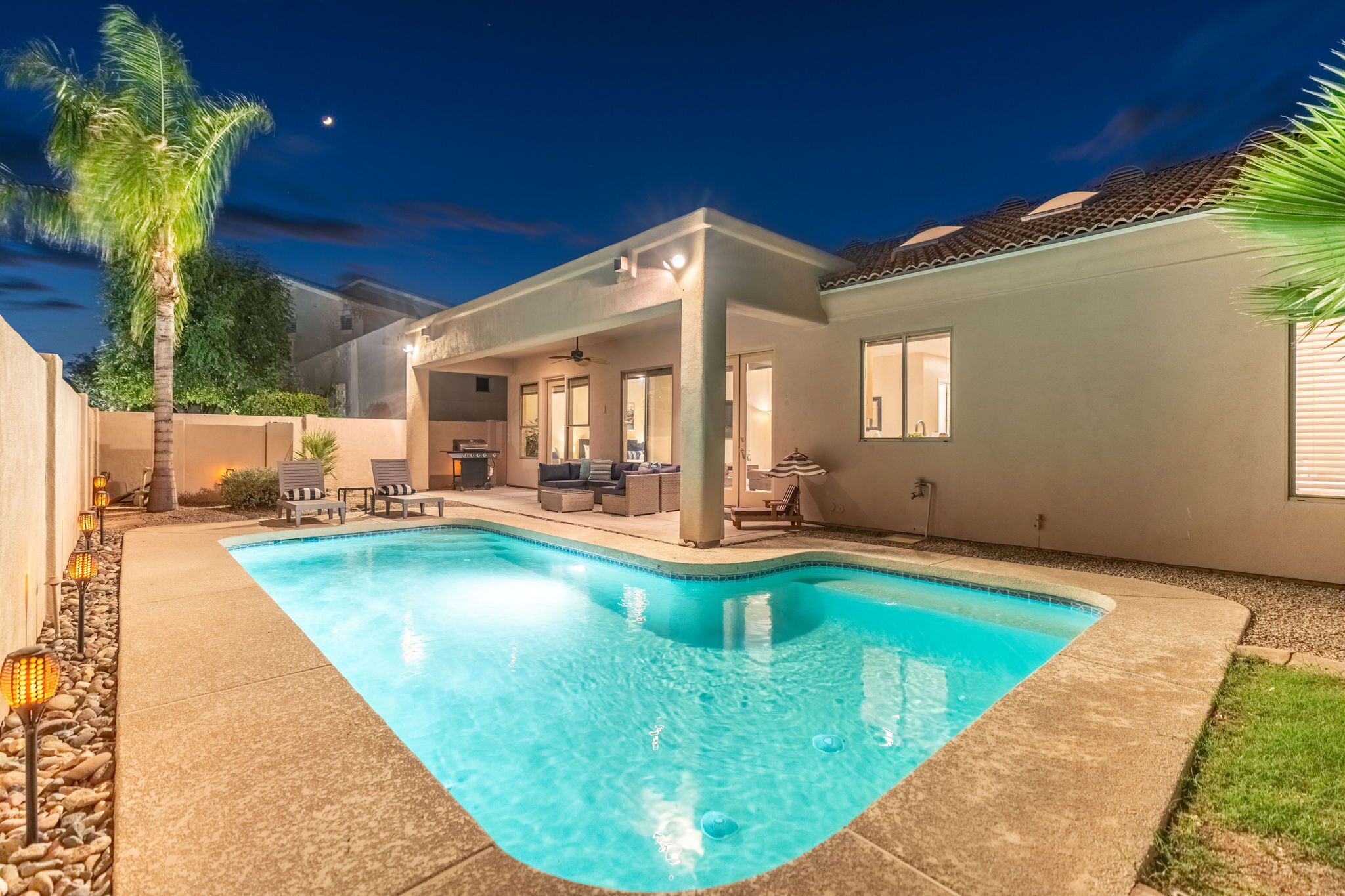 A residential backyard with a swimming pool, outdoor seating, and a covered patio area.