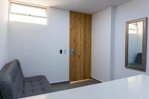Una habitación con puerta de madera, un sofá gris, una mesa blanca y un espejo con marco decorativo.