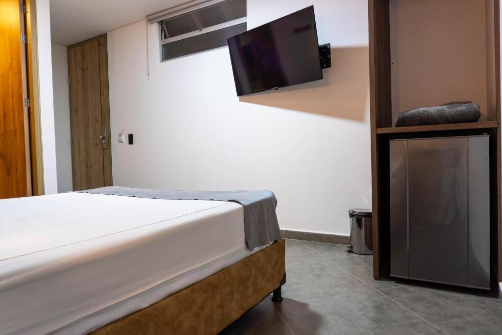 Une chambre à coucher avec un lit, un téléviseur fixé au mur, une petite armoire et une poubelle.