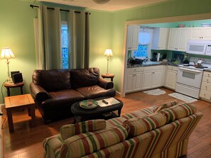 Un salon avec un canapé en cuir marron, un divan à rayures et une petite table avec une lampe. Il y a également un coin cuisine avec des placards blancs et un four à micro-ondes.