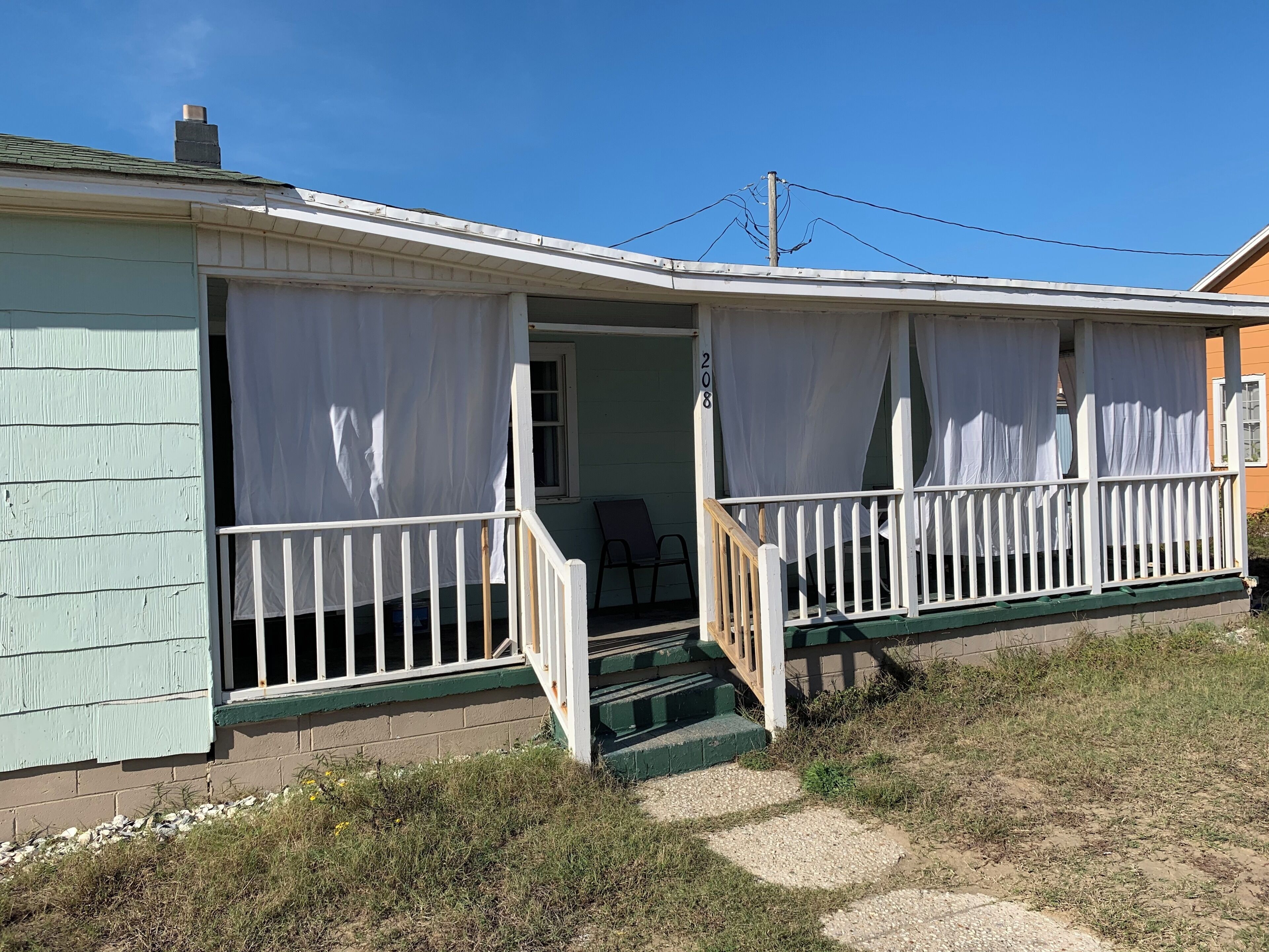 A single-story house with a porch, white curtains, and a number '208' on the wall.