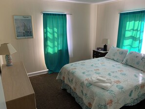 Une chambre d’hôtel avec un lit, une table de chevet, une lampe, un tableau encadré et deux fenêtres avec des rideaux.