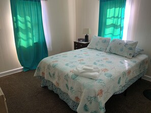 Ein ordentlich bezogenes Bett mit floralem Bettzeug, ein Nachttisch mit Lampe und Vorhänge.