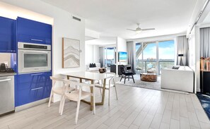 Ein modernes Hotelzimmer mit Essbereich, einer Küche mit blauen Schränken, einem großen Fenster mit Stadtblick und einem Deckenventilator.