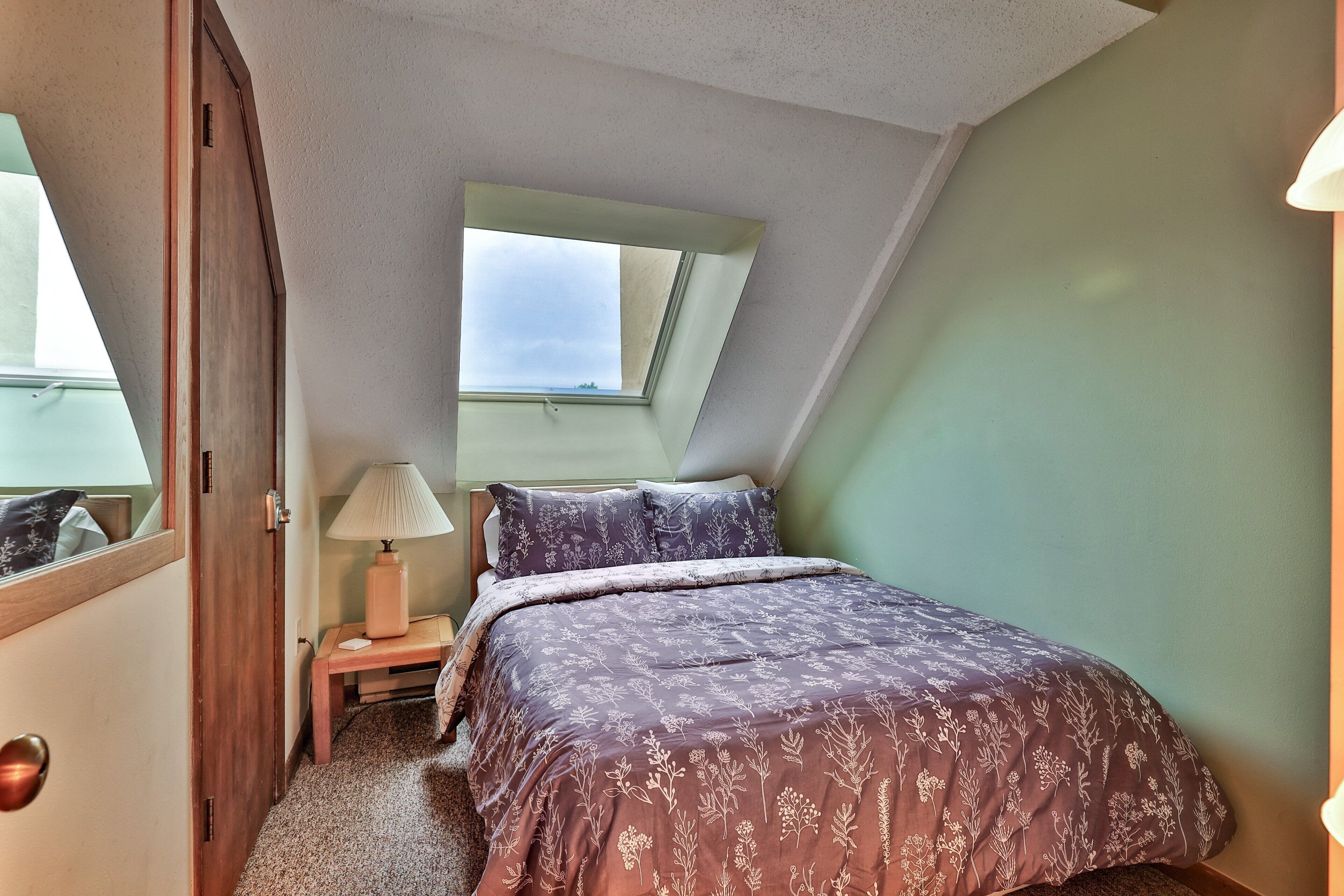 A bedroom with a sloped ceiling, a skylight, a bed with floral bedding, a bedside table with a lamp, and a door.