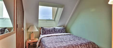 A bedroom with a sloped ceiling, a skylight, a bed with floral bedding, a bedside table with a lamp, and a door.