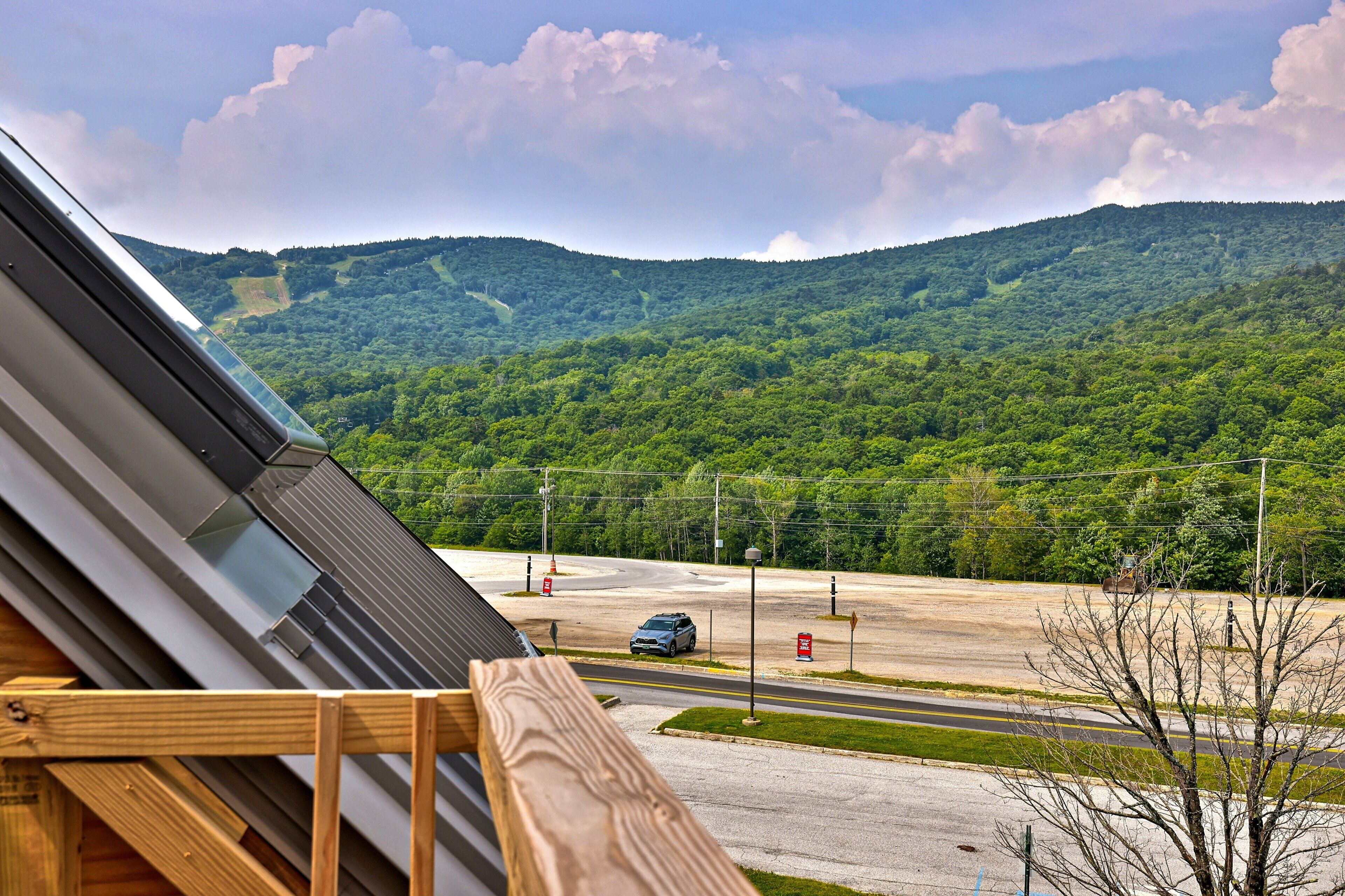 A view from a window overlooking a road with a car, a parking lot, and a forested mountain range.