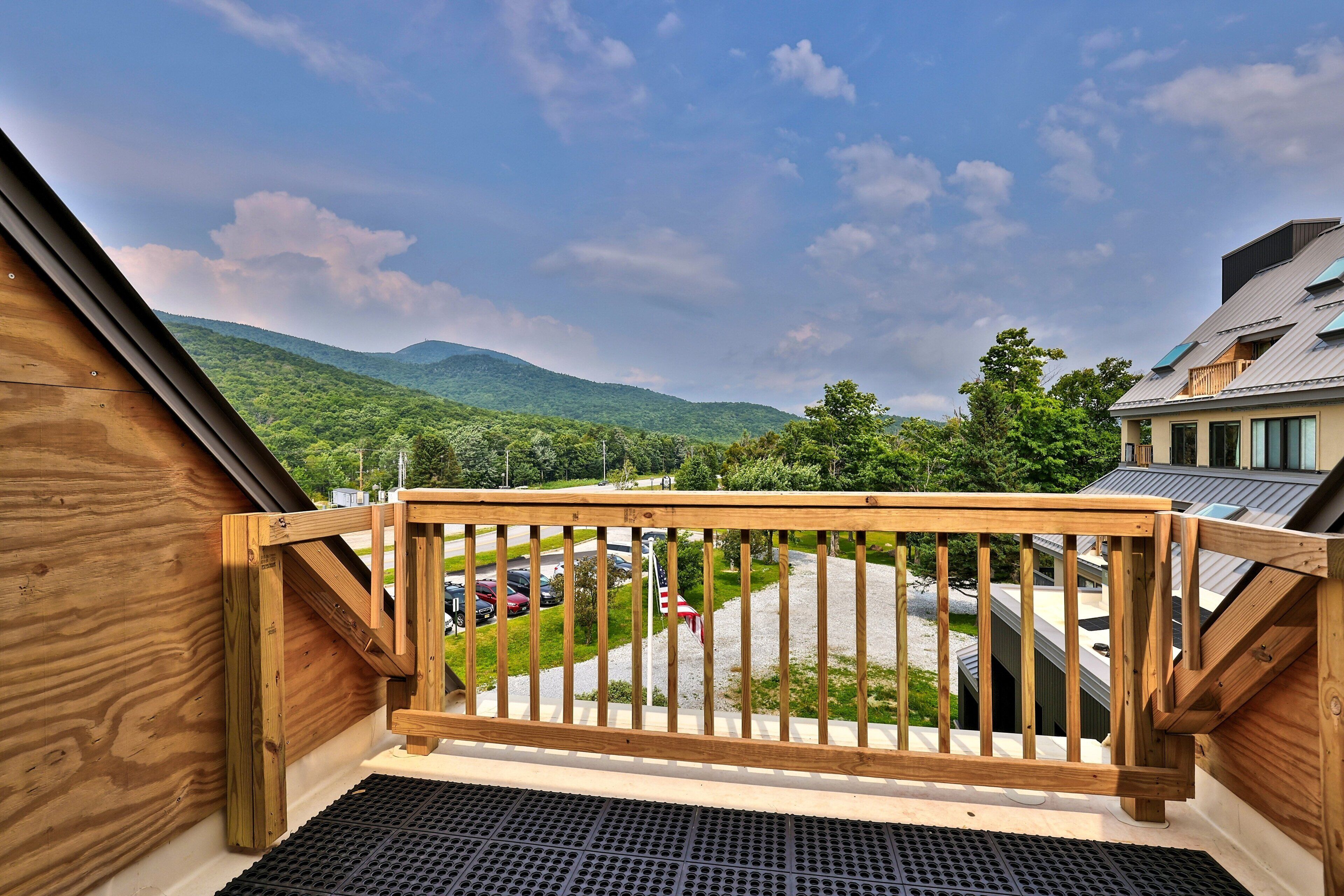 A wooden balcony with a view of a parking area and green hills in the distance.