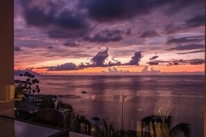 A panoramic view of a coastal landscape with a sunset, featuring a balcony with wicker furniture and palm trees.