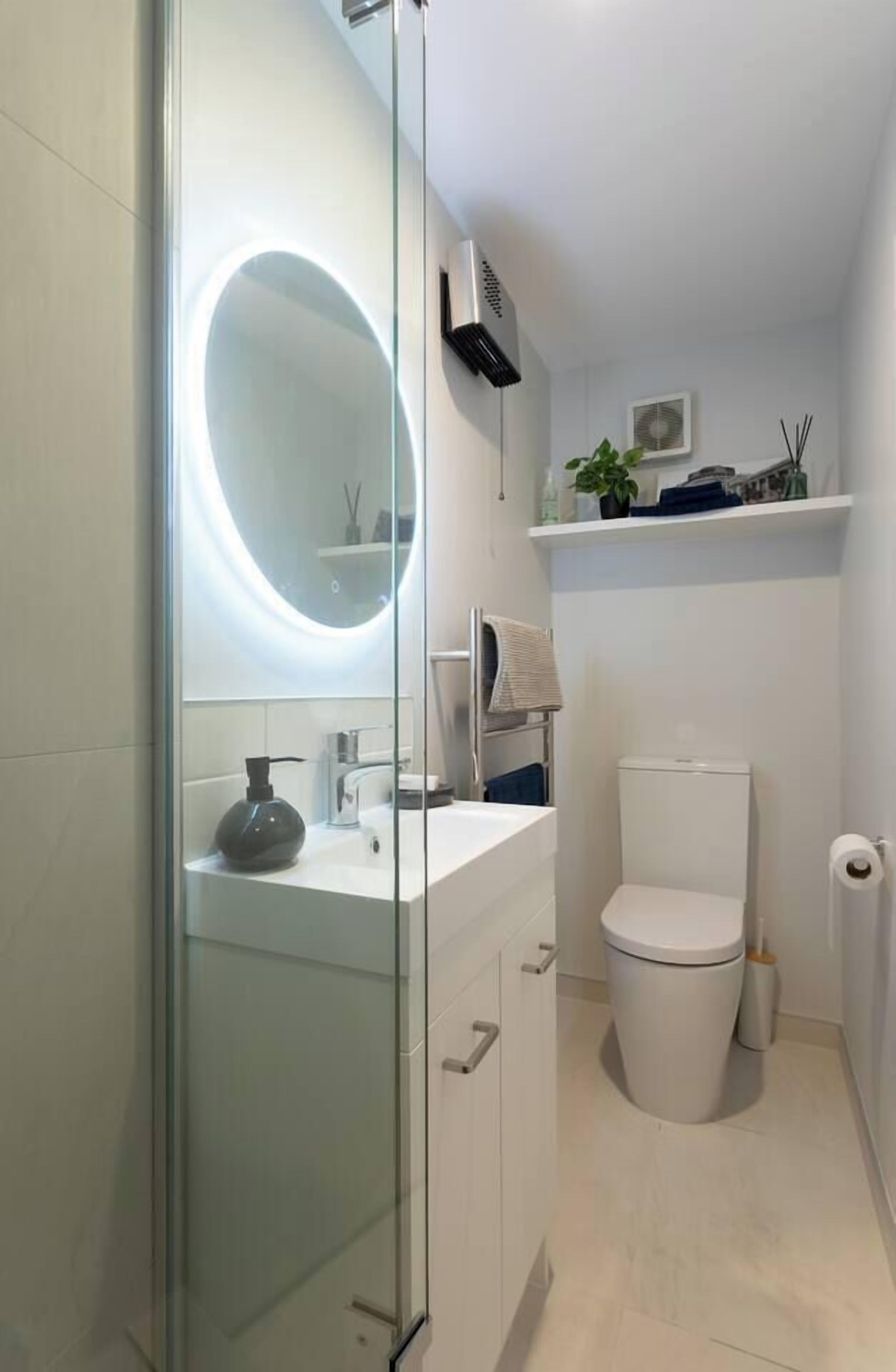 A modern bathroom with a glass-enclosed shower, a round mirror with built-in lighting, a white toilet, and a small shelf with toiletries.