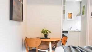 Quarto pequeno e acolhedor com uma mesa de jantar de madeira, duas cadeiras, um vaso de planta, uma cama com cobertor cinzento e uma área de cozinha visível por uma porta aberta.