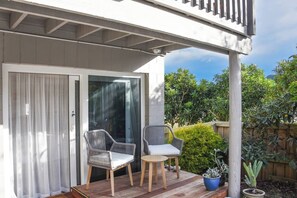 A covered patio with two chairs and a small table, a sliding glass door, and a view of greenery.