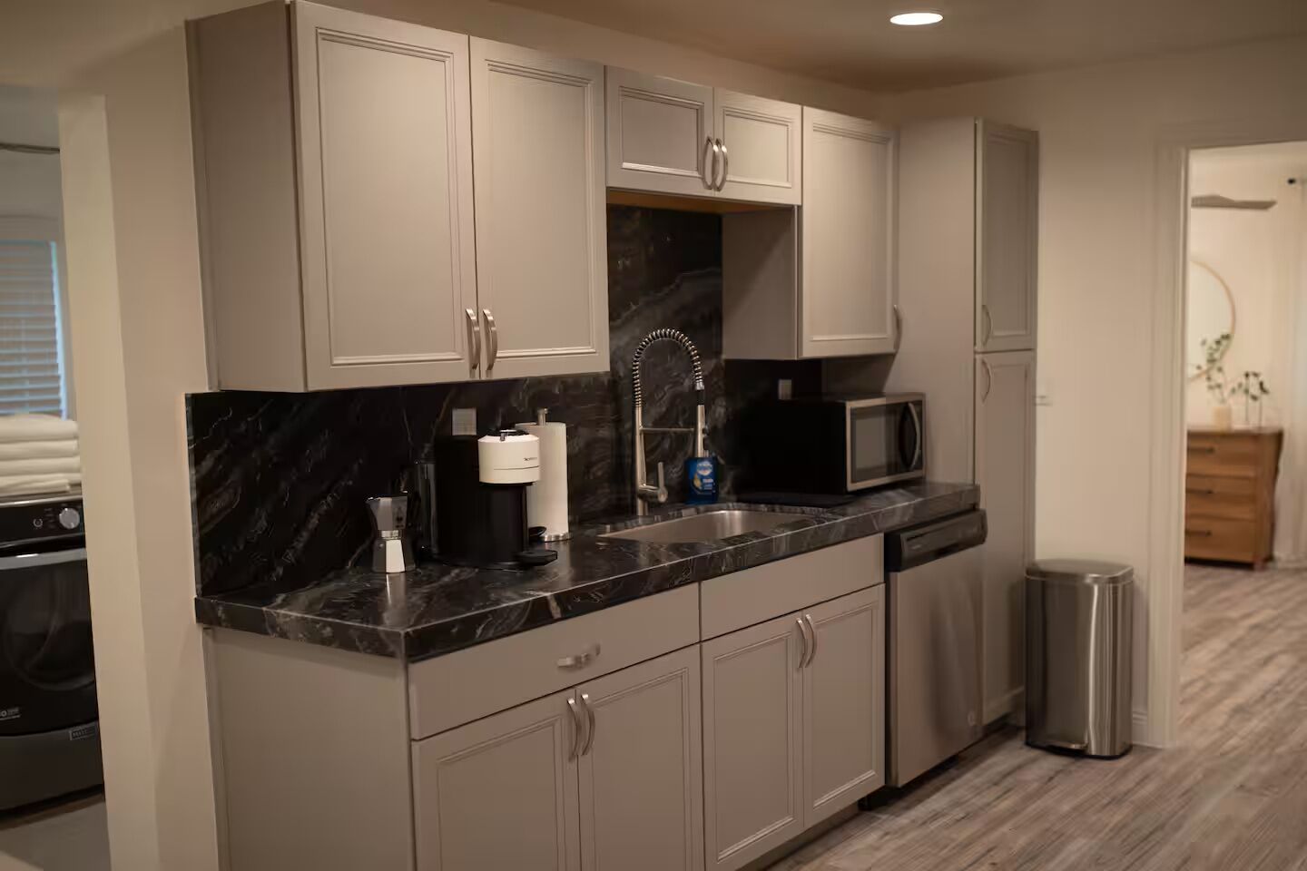 A modern kitchen with grey cabinets, a black marble countertop, a stainless steel sink, a coffee maker, a microwave, and a dishwasher.
