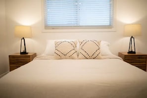 A bed with two heart-shaped pillows, a window with blinds, and two bedside lamps on wooden nightstands. - Sunnyside A - Spacious 3-bedroom house in peaceful Clovis with AC (Clovis)