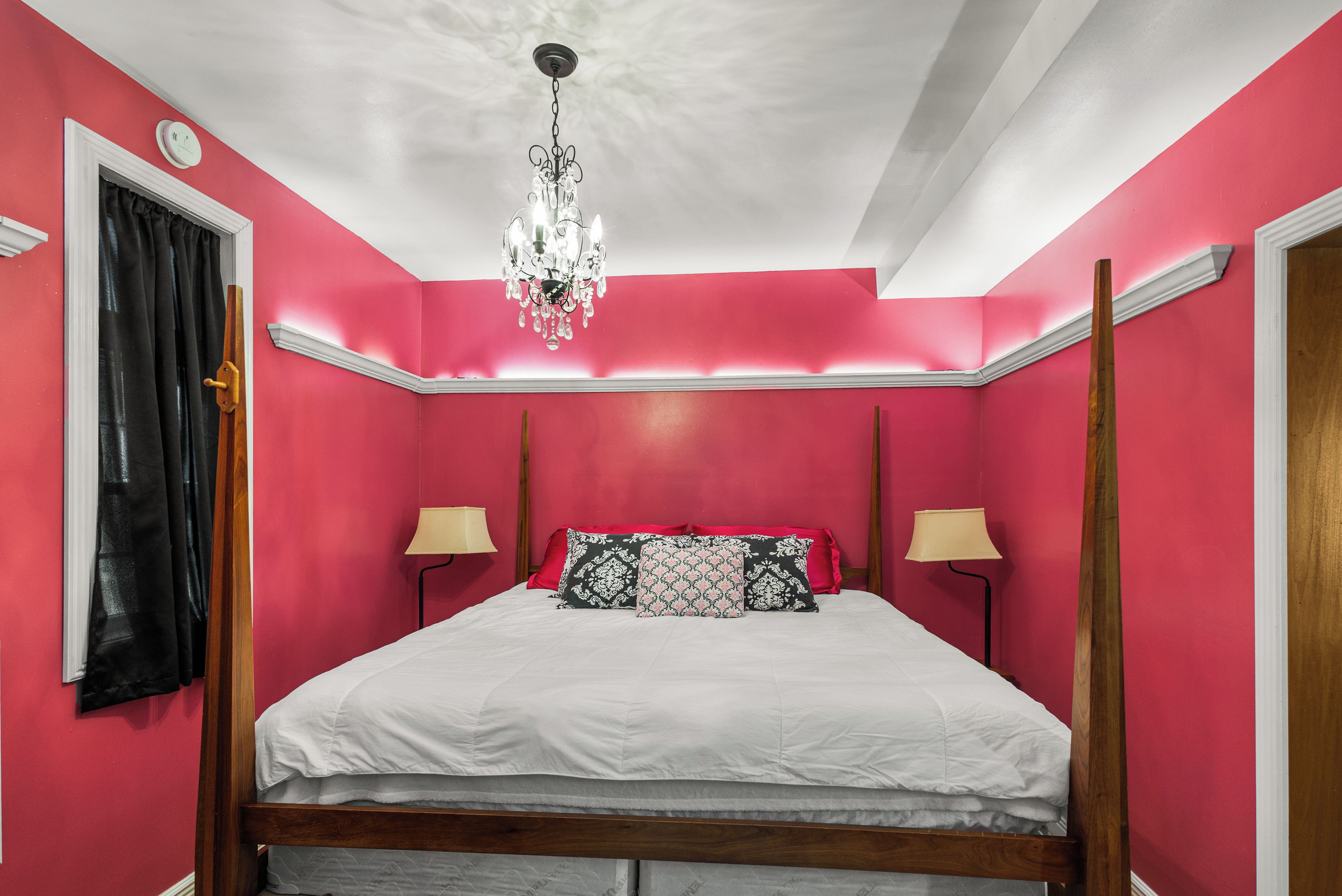 A bedroom with a four-poster bed, two bedside lamps, a chandelier, and a window with curtains.