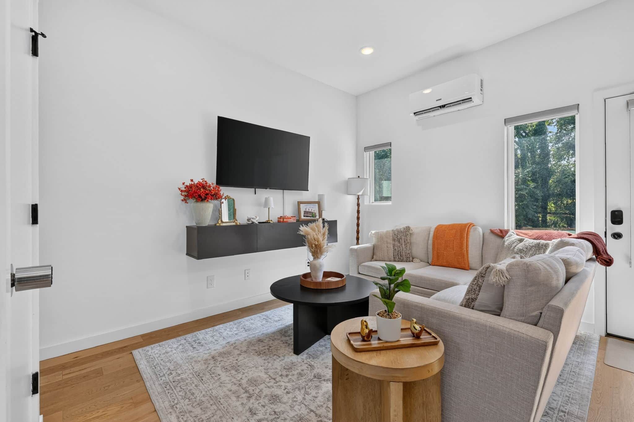 A modern living room with a sofa, a coffee table, a flat-screen TV mounted on the wall, and a window with a view of trees.