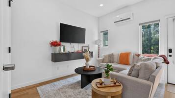 A modern living room with a sofa, a coffee table, a flat-screen TV mounted on the wall, and a window with a view of trees.