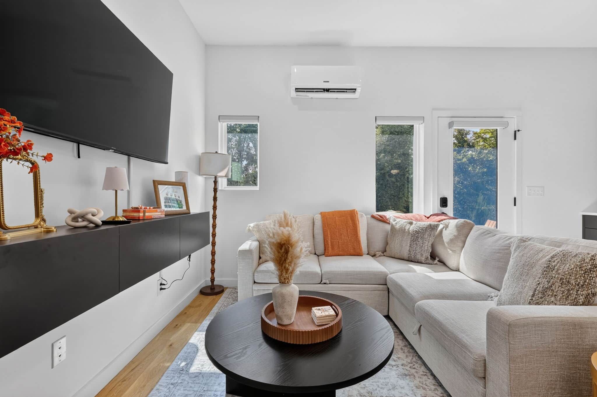A modern living room with a sectional sofa, a flat-screen TV mounted on the wall, and a large window providing natural light.