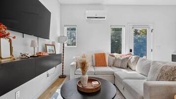 A modern living room with a sectional sofa, a flat-screen TV mounted on the wall, and a large window providing natural light.
