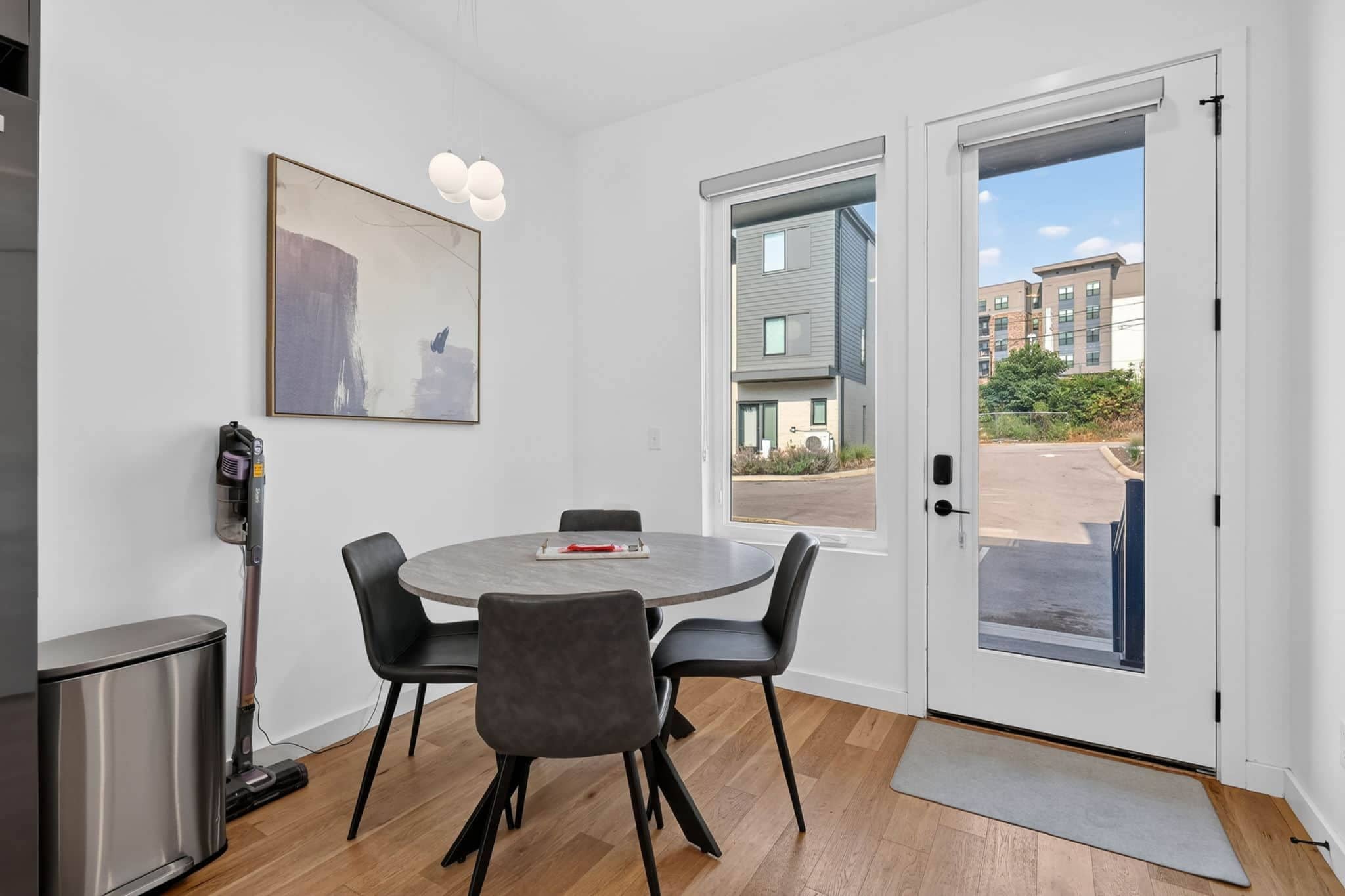 Une pièce moderne avec une table ronde, quatre chaises, un tableau, un aspirateur et une porte donnant sur l’extérieur.
