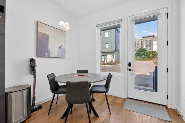 A modern room with a round table, four chairs, a painting, a vacuum cleaner, and a door leading outside.