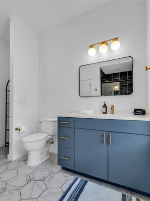 Un baño moderno con un inodoro blanco, un tocador con un armario azul oscuro y un espejo con tres luces.