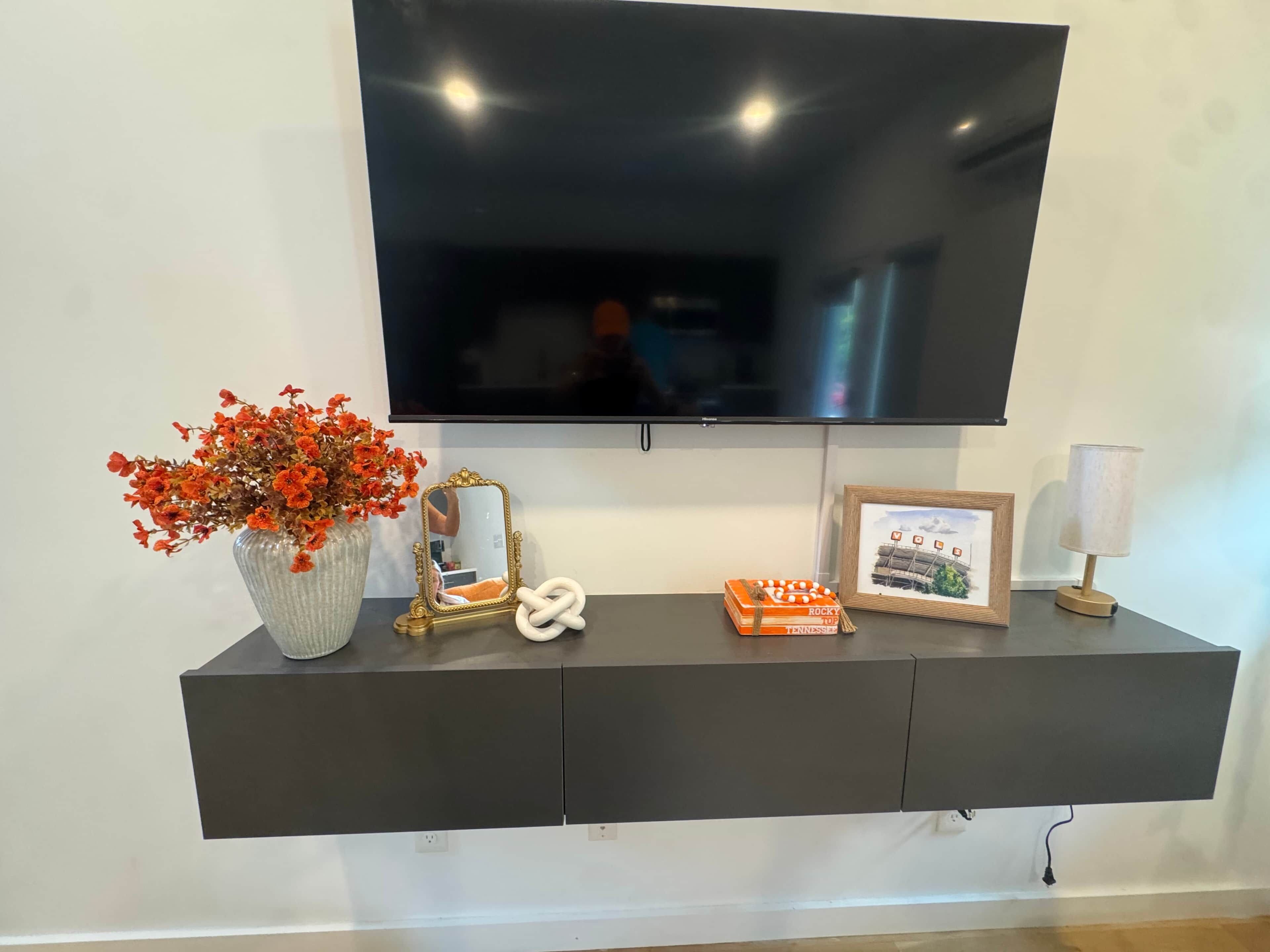 A flat-screen TV mounted on a dark wall above a black console with a vase of flowers, a framed picture, and a small lamp.