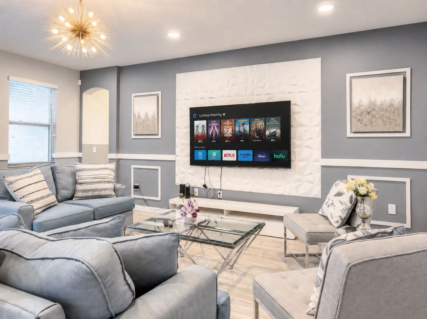 Ein modernes Wohnzimmer mit grauen Sofas, einem Glastisch und einem flachbildfernseher an der Wand, der verschiedene Streaming-Dienste anzeigt.