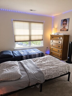 Quarto com duas camas, cômoda, janela com persianas e luminária fixada na parede.
