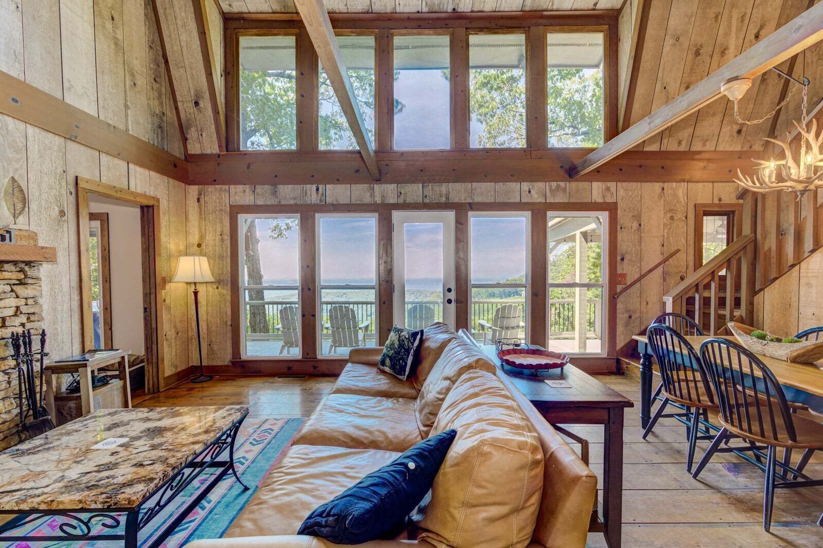 A spacious room with a large window, a wooden ceiling, a sofa, a dining table, and a chandelier.
