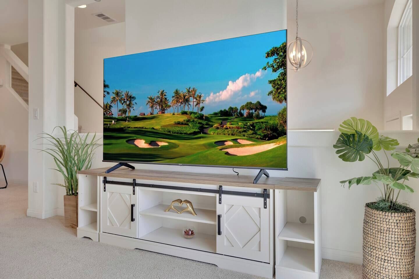 Un televisor de pantalla plana montado en un mueble blanco con una imagen de un campo de golf, una lámpara colgante y plantas en macetas.