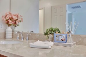 Une salle de bain avec un lavabo, deux robinets, un miroir, un vase avec des fleurs, une plante et un distributeur de savon.