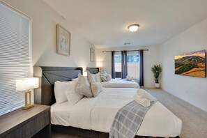 Une chambre d’hôtel avec deux lits, une table de chevet avec une lampe, une fenêtre avec des stores, un tableau au mur et un luminaire au plafond.