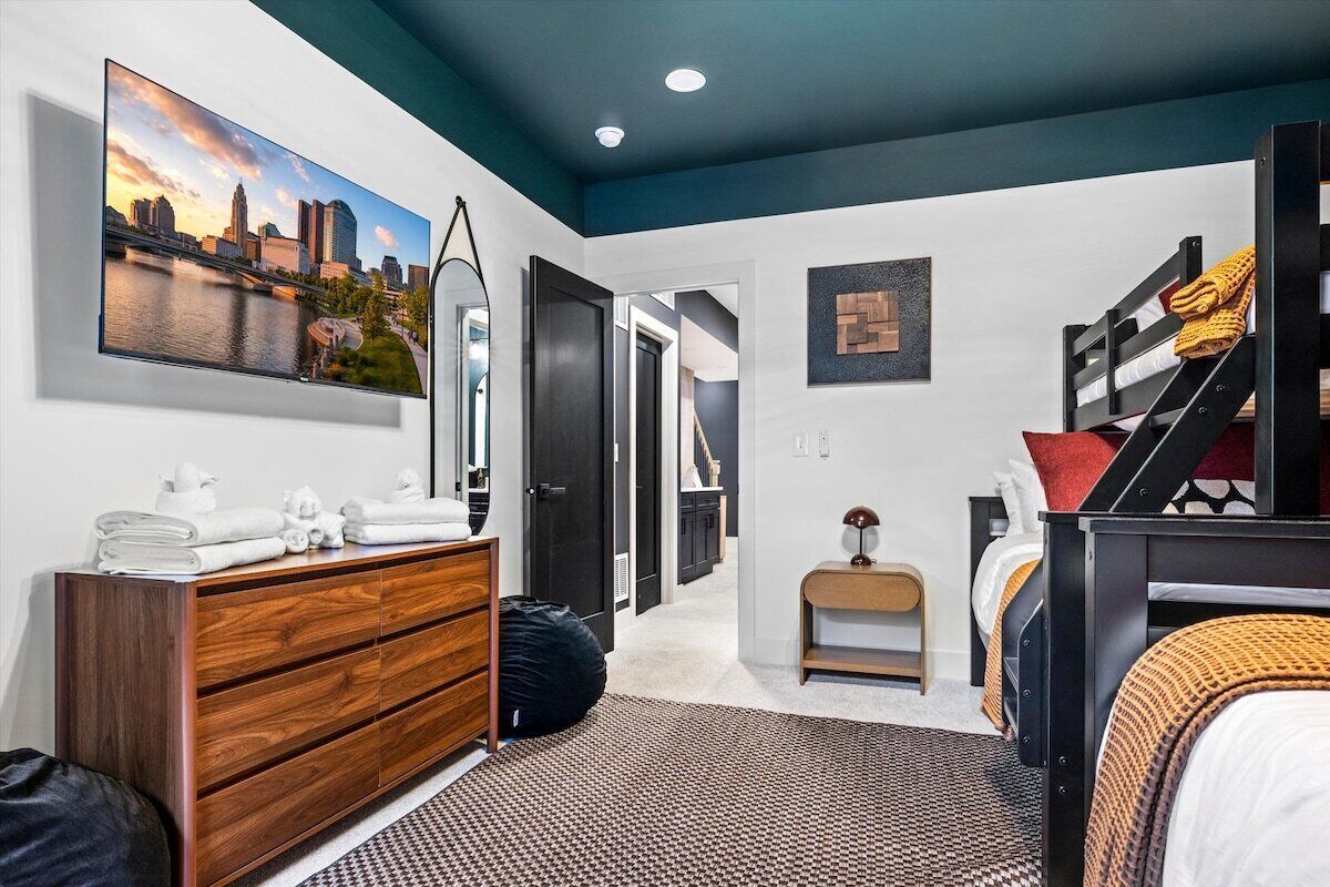 A bedroom with a cityscape painting, a wooden dresser, a bed with a patterned bedspread, and a staircase.