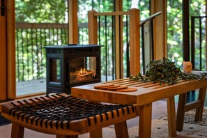 A cozy room with a wood-burning stove, a woven chair, and a table with wooden utensils and a plant. - Treeangle Treehouse - Romantic Treehouse for 2 | Firepit | Hot Tub (Morganton)