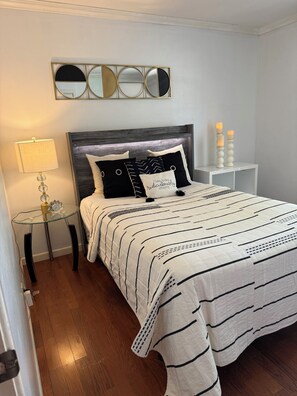 Een net opgemaakt bed met een gestreept dekbed, een nachtkastje met een lamp en een spiegel aan de muur.